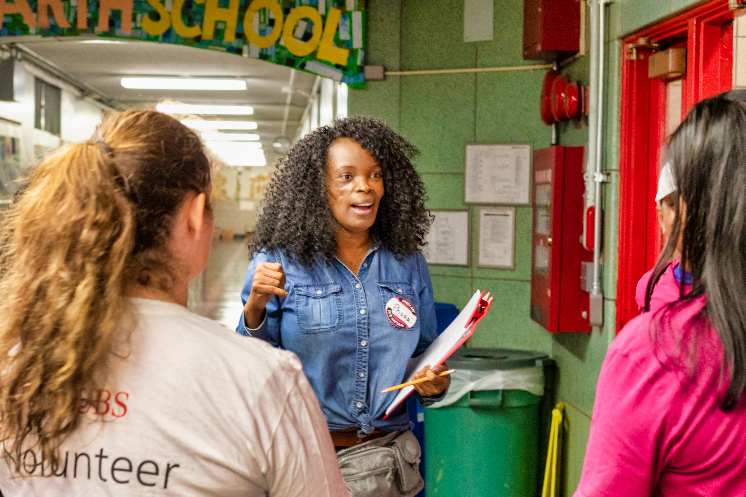 A woman with a clipboard speaks to two volunteers in a school hallway