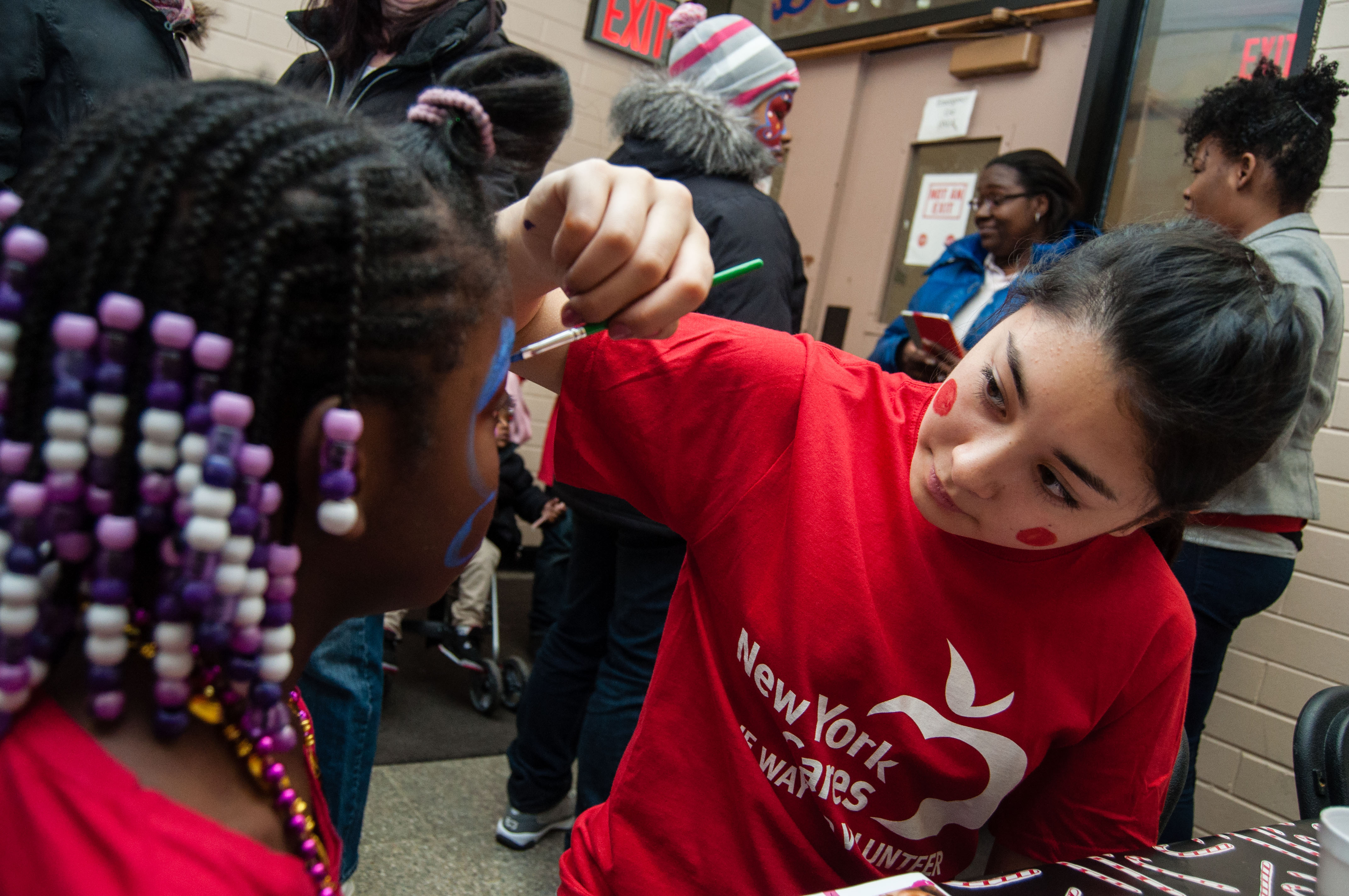 A teenage girl in a red New York Cares shirt applies face paint to a child's face