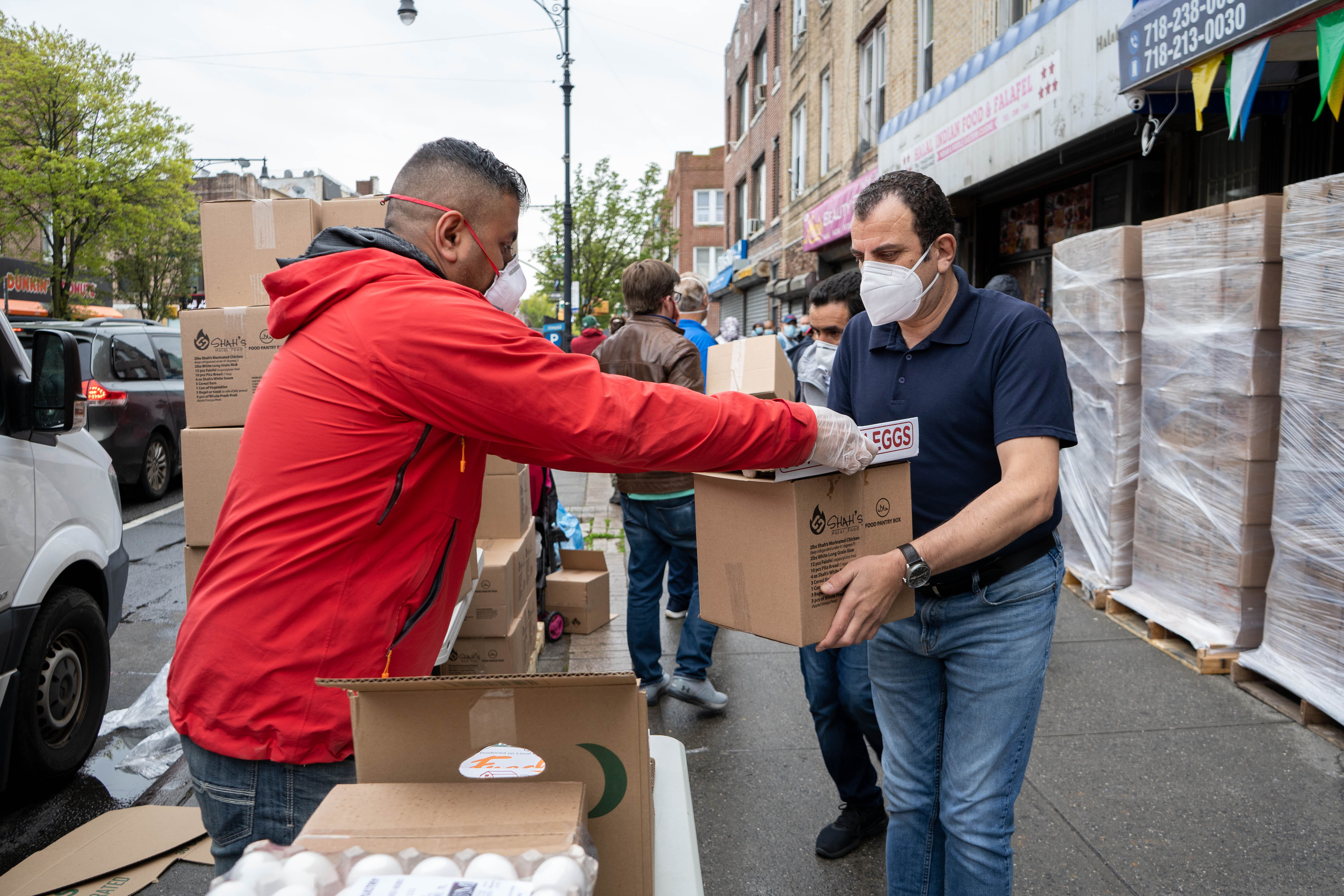 A man in a red jacket passes a box of food supplies to another man at a sidewalk food pantry